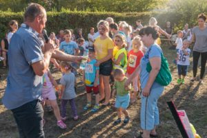 Mężczyzna z mikrofonem i butelką wody w dłoni. Przed nim stoi grupa dzieci z pomalowanymi twarzami.