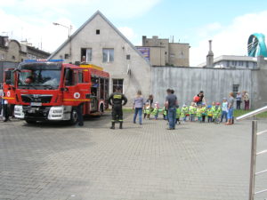 Duży plac na którym od lewej stoi wóz strażacki, na środku strażak przeprowadzający pokaz dla grupy dzieci i kilkoro rodziców po prawej.