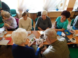 Grupa seniorów siedzi przy długim stole, " Seniorzy wymieniają się technikami zdobienia kartek świątecznych."