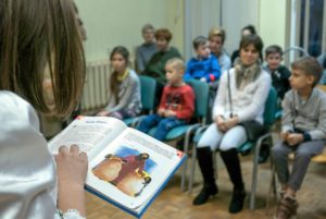 Na pierwszym planie otwarta książka w rękach czytającej dziewczynki, w tle dzieci i rodzice na widowni